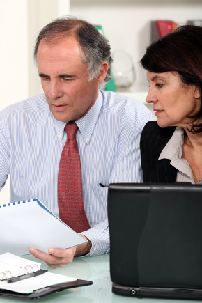 Man in striped shirt holding spiral binder and talking to woman at laptop