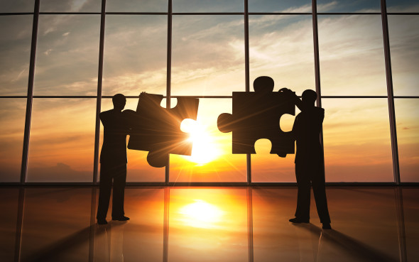 Two businessmen holding giant puzzle pieces in silhouette against a sunrise