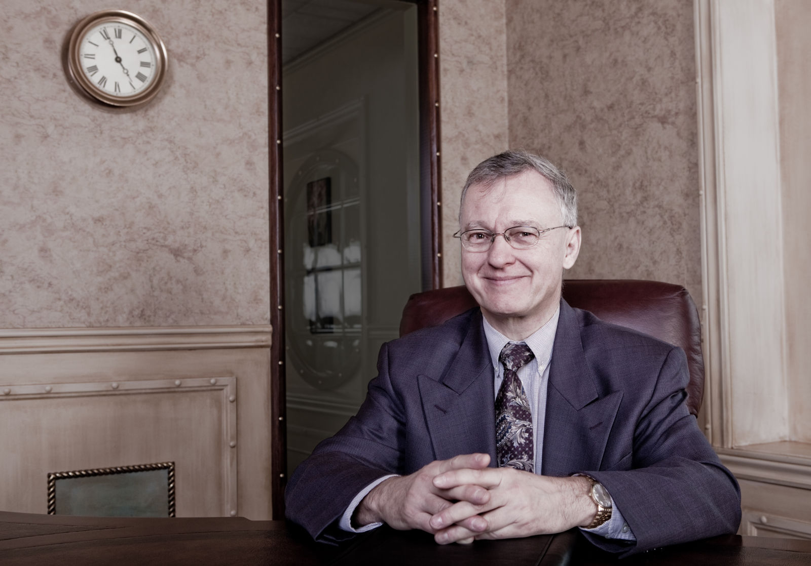 Older businessman sitting at desk smiling as wall clock indicates 5 minutes to retirement