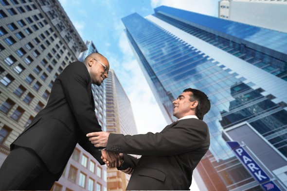 Two businessmen shaking hands in front of tall office buildings