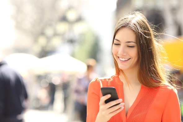 Woman wearing orange shirt texting on a smartphone walking in the street in a sunny day