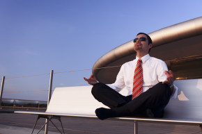 Calm balanced businessman sitting outdoors on bench in Yoga lotus pose meditating, with office building and blue sky in background