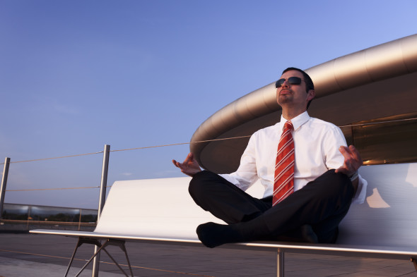 Calm balanced businessman sitting outdoors on bench in Yoga lotus pose meditating, with office building and blue sky in background