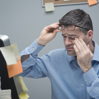 Man rubbing his eye and holding glasses while looking at computer with notes stuck to side