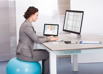 Businesswoman with both desktop and laptop computers, sitting on Pilates ball