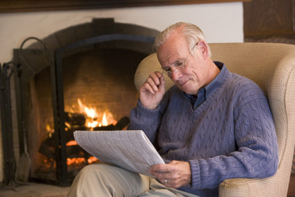 Older man reading newspaper by fireplace
