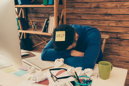 Man with head down on desk, sticky note with "HELP" writte on it on baseball cap