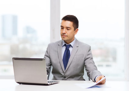 Man holding papers and working on laptop in front of large window