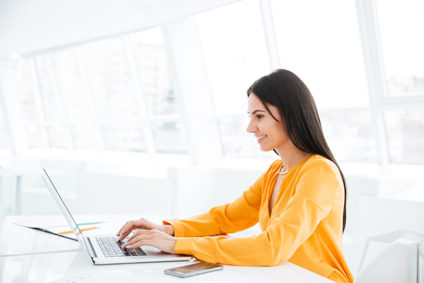 Businesswoman in orange shirt using laptop computer by white wall of windows