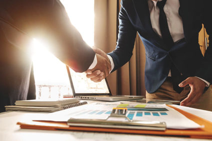 Two businessmen shaking hands across desk and documents in office